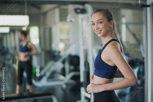 Young sport Caucasian woman shot hair is measuring waist in gym near big mirror. Slim woman use measuring yellow tap and looking at camera in sport club. Woman felling happy and smiling.
