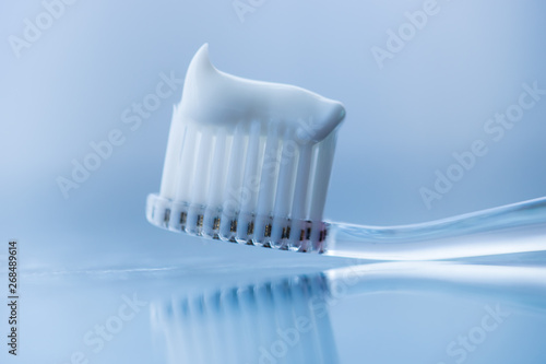 Transparent plastic toothbrush with white toothpaste on a blue white background with reflection on the glass.