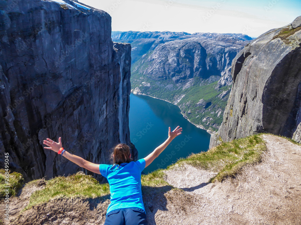 Obraz premium A girl lying at the edge of a steep mountain pretends to be flying. She spreads arms wide, in a gesture of freedom. In front of her stunning Lysefjorden shimmering with many shades of blue and green