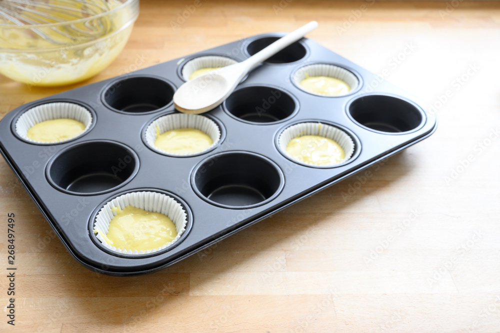 muffin tin, half filled with paper cups and dough for cupcake baking on a wooden kitchen table, copy space muffin tin, half filled with paper cups and dough for cupcake baking on a wooden kitchen table, copy space