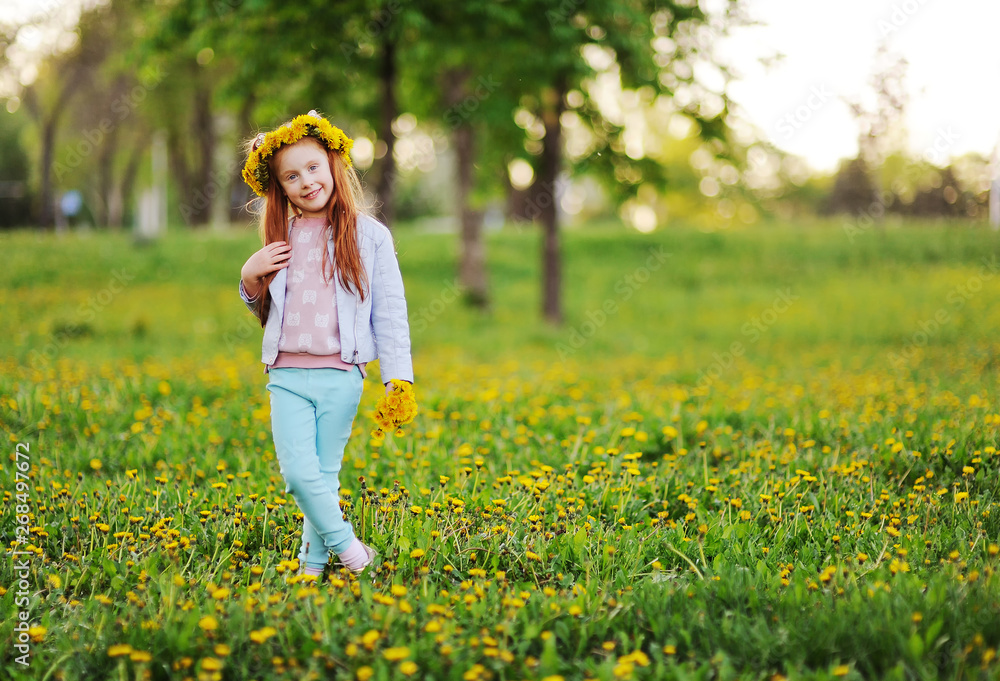 Fototapeta premium a little girl with red hair smiles against a field of dandelions and green grass. Summer, childhood, holidays. space for text, copy space,