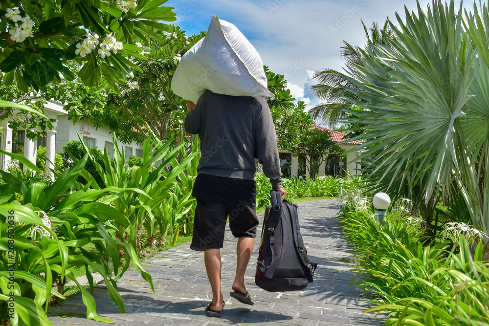 A man in a gray sweater and black shorts carries a white bag on his shoulder and holds a backpack in his hand. He walks along a stone path among palm trees and green plants around the hotel