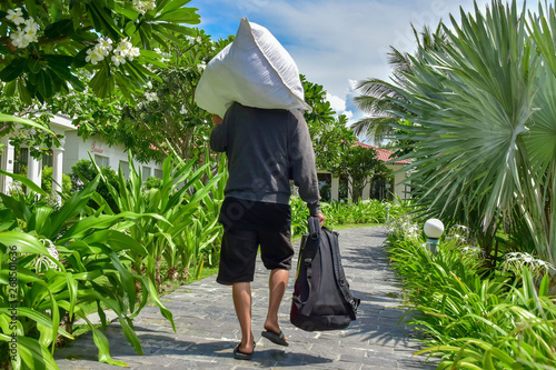 A man in a gray sweater and black shorts carries a white bag on his shoulder and holds a backpack in his hand. He walks along a stone path among palm trees and green plants around the hotel