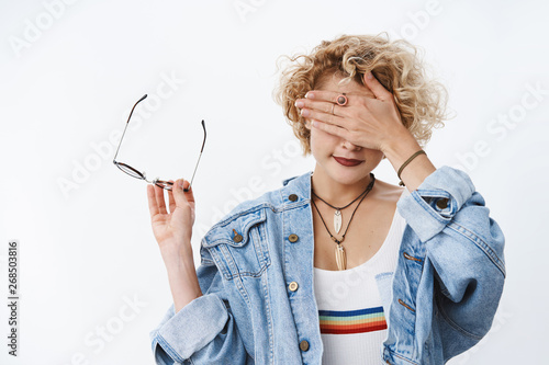Tell me when it over. Portrait of girl cannot look at gross thing taking off glasses holding frames in hand as covering sight with palm and smiling waiting for time to open eyes over white wall