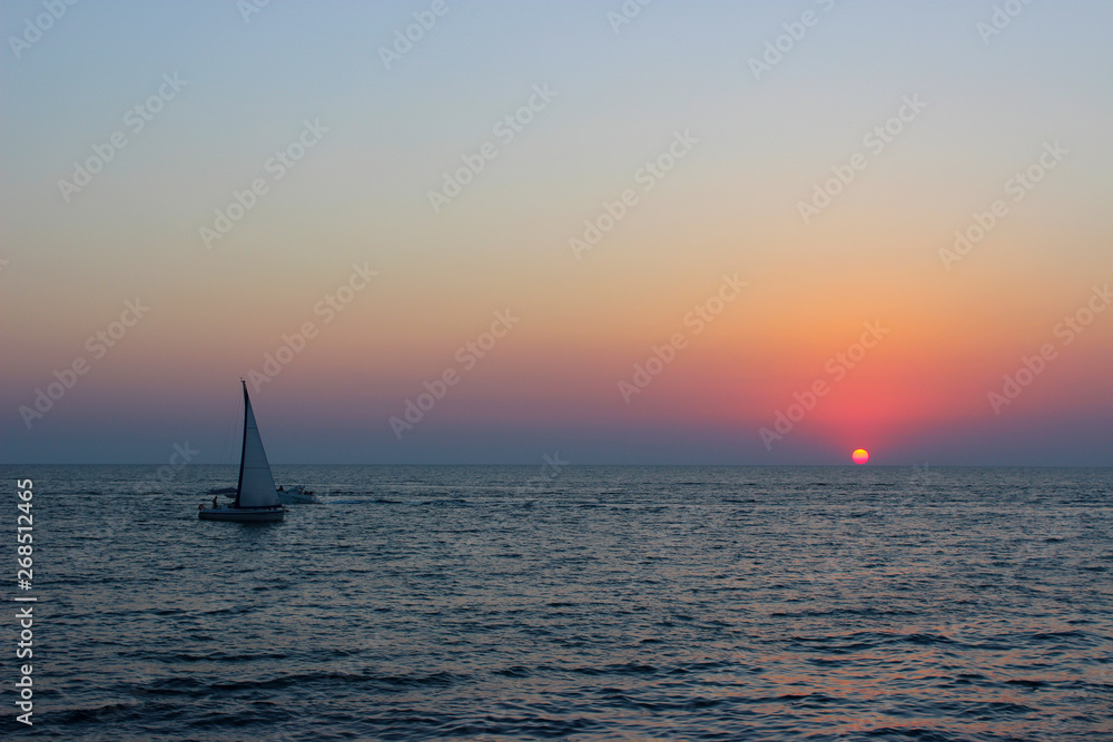 Sea sunset. The sun is very low. Sky and water in red and blue tones. Small waves. On the horizon, a small sailboat. Warm summer.