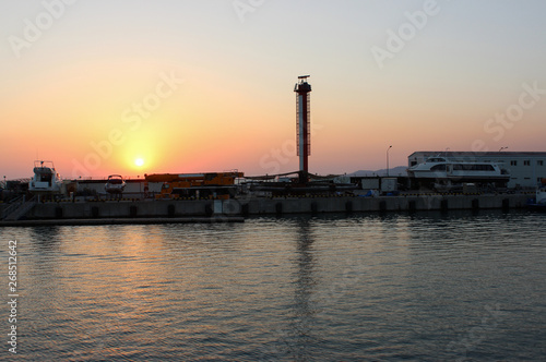 Sunset on sea. The sun painted the sky and water red and yellow. In the distance the dark line of the pier dock. It shows the silhouettes of boats, vessels, crane, tower. Summer is warm, the horizon.