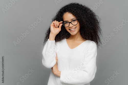 Portrait of happy woman with curly hair, kepes hands on frame of glasses, has gentle smile, wears white jumper, isolated over grey background. Copy space for your advert. Delighted student indoor