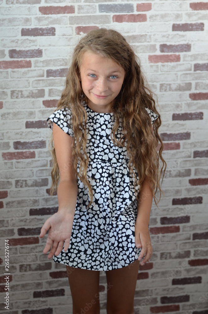 Little girl with blue eyes in a short black and white dress with hearts and a hat by the wall
