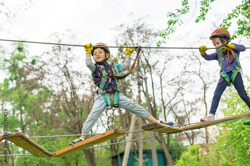 Two cute happy young children, boy and girl in protective harness, carbine and safety helmets on rope way on bright sunny day on green foliage bokeh background. Outdoors activities, games concept.
