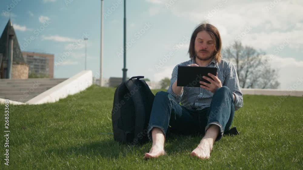 Handsome man working with tablet in park in autumn. Young man sitting on the green grass reading tablet ipad after work Handsome man working with tablet in park in autumn. Young man sitting on the green grass reading tablet ipad after work