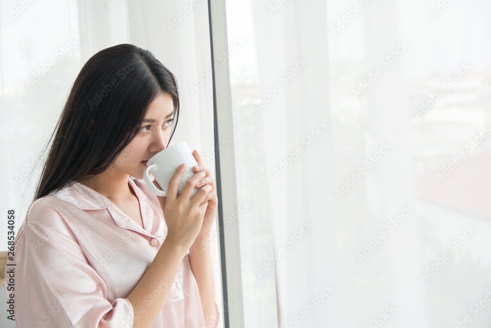 Side view portrait a pretty woman thinking looking holding a coffee mug beside a window at home Side view portrait a pretty woman thinking looking holding a coffee mug beside a window at home