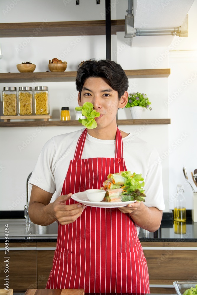 Young good looking man in red apron presenting the healthy menu of salad roll with smile, prepared in home kitchen Young good looking man in red apron presenting the healthy menu of salad roll with smile, prepared in home kitchen