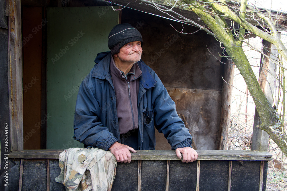 An old man in sloppy clothes stands on the threshold of his own ruined house and looks into the distance An old man in sloppy clothes stands on the threshold of his own ruined house and looks into the distance