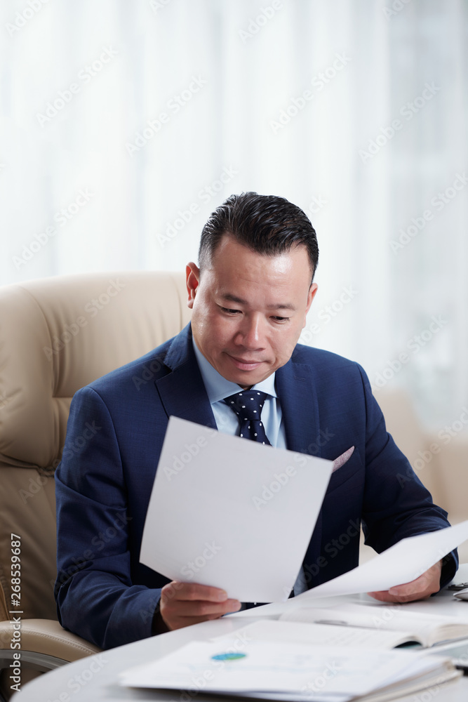 Asian mature businessman sitting at the workplace and working with documents, he reading and examining business contract during his work day at office Asian mature businessman sitting at the workplace and working with documents, he reading and examining business contract during his work day at office