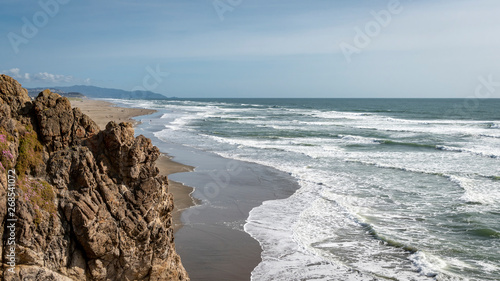 Spring afternoon at Ocean Beach in San Francisco.  High angle shot looking south from Cliff House.  Mild surf, people enjoying a relatively warm day.  Blue sky.  Rough cliffs foreground.
