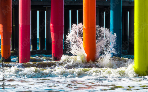 Fotografia Wave brakes into orange colorful pier footing lit up with a sun with other rainb