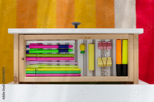 pens, pencils, markers, felt-tip pens, erasers, clips of bright orange, yellow, pink, green colors are neatly folded in size in a white table drawer against a wooden gray floor
