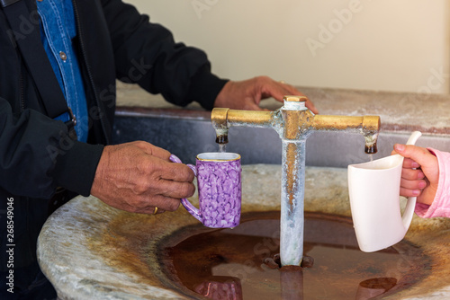 Fotografia Closeup view of hands hold beautiful ceramic glass souvenirs and filling mineral springs water from golden faucet at Army spa institution in Spa Town, Bohemia Region, Karlovy Vary, Czech Republic