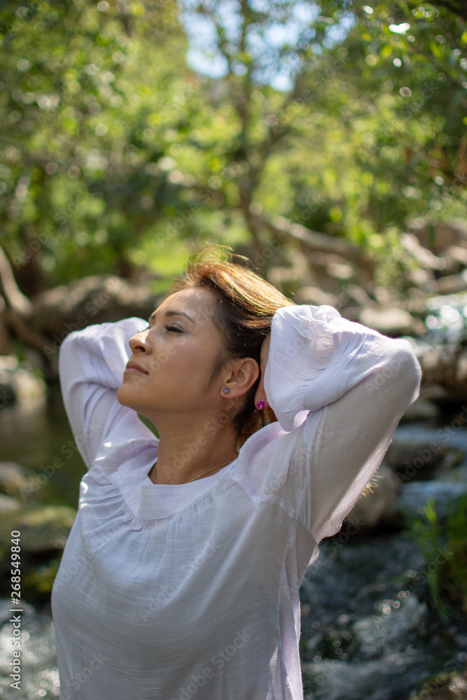 Latina woman throwing her hair back with her hair in the sun in front of woods and a stream in the shade