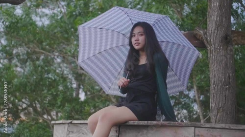 Slow motion footage of girl sitting on deck holding an umbrella. The girl dangles her legs and looks toward camera with green leaves and a tree trunk visible behind her. photo