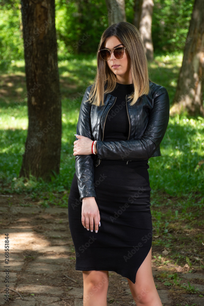 © Emilija - Beautiful fashion woman model in black dress and leather jacket and sunglasses posing in nature on a sunny spring day. Forest in background. Fashion concept. Close up, selective focus © Emilija - Beautiful fashion woman model in black dress and leather jacket and sunglasses posing in nature on a sunny spring day. Forest in background. Fashion concept. Close up, selective focus