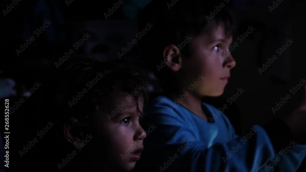 Boy playing console video games with his toddler brother on a high definition television screen reflected on their face together with the excitment of playing digital electronic games. Boy playing console video games with his toddler brother on a high definition television screen reflected on their face together with the excitment of playing digital electronic games.
