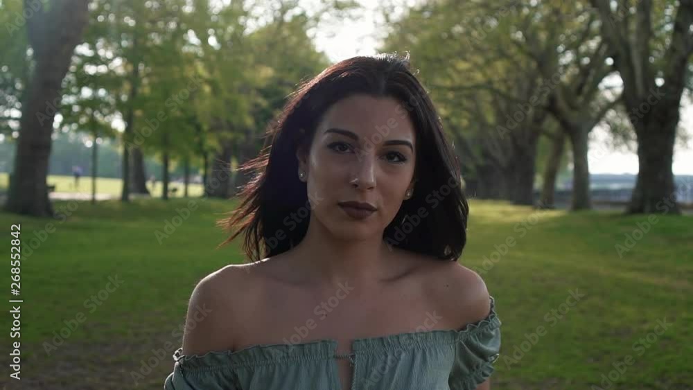 Slow Motion Portrait of a gorgeous hispanic latino young woman looking at the camera and show emotions from serious to smiling and laughing with a beautiful British park in the background. Slow Motion Portrait of a gorgeous hispanic latino young woman looking at the camera and show emotions from serious to smiling and laughing with a beautiful British park in the background.