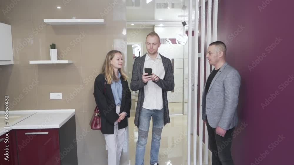 The splendid coule talks with the real estate agent in the modern kitchen. They all observe new apartment that they would like to buy. Tall guy raises his hand with the phone and tries to take photos The splendid coule talks with the real estate agent in the modern kitchen. They all observe new apartment that they would like to buy. Tall guy raises his hand with the phone and tries to take photos