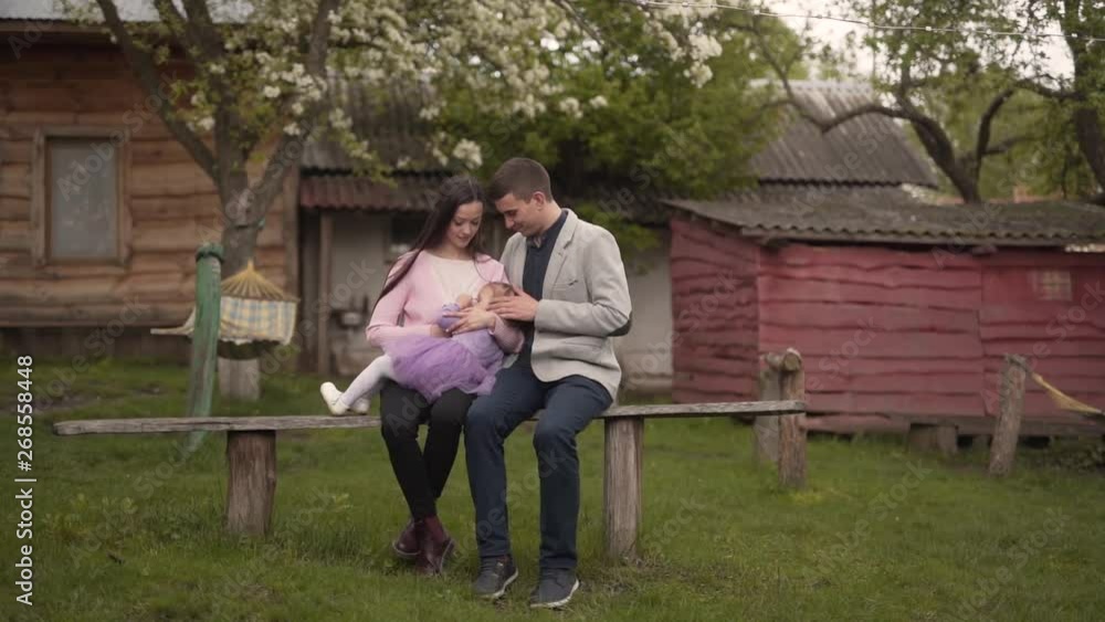 Young Caucasian Family Sitting on Bench in Countryside Village Backyard With Wooden Houses While Mother Breastfeeding Their Baby Girl. Light Camera Movement Young Caucasian Family Sitting on Bench in Countryside Village Backyard With Wooden Houses While Mother Breastfeeding Their Baby Girl. Light Camera Movement