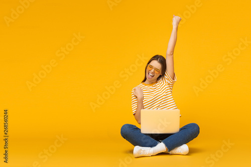 Winner! Excited smiling girl sitting on floor with laptop, raising one hand in the air is she wins, isolated on yellow background photo