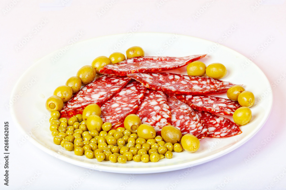 On the plate lies sliced sausage, green peas, and olives. On the plate lies sliced sausage, green peas, and olives.