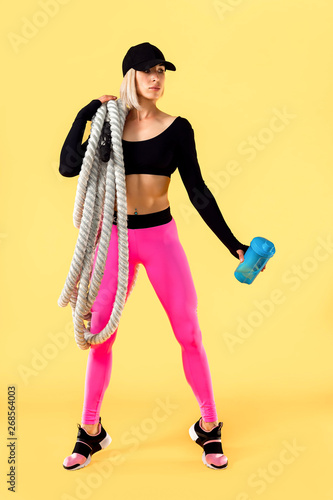 Attractive woman in pink and black sportswear with heavy ropes on her shoulders and holding shaker on yellow background. Strength and motivation. Sporty woman working with heavy ropes.