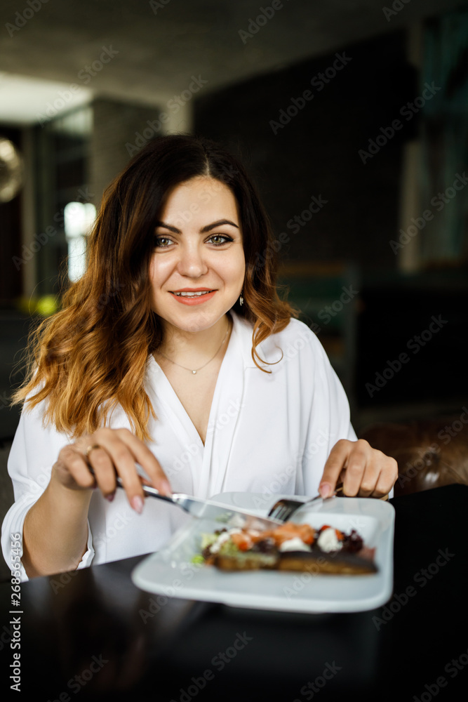 A young, sympathetic woman, not a thin-headed body building, sits in a cozy cafe and eats a salad. Business clothing style.