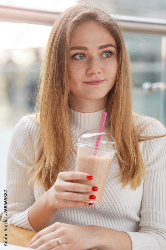 Close up portrait of pensive young blonde student in white sweater and long hair, sitting in cafe, holds plastic cup with fresh beverage, attractive woman drinks cocktail and looks thoughtfuly aside. Close up portrait of pensive young blonde student in white sweater and long hair, sitting in cafe, holds plastic cup with fresh beverage, attractive woman drinks cocktail and looks thoughtfuly aside.