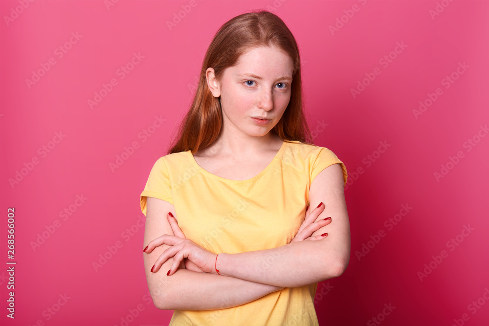 Studio shot of serious harsh girl looking questioningly directly at camera, keeps arms crossed, dressed yellow casual t shirt, isolated over pink background. Copy space for advertisment or promotion. Studio shot of serious harsh girl looking questioningly directly at camera, keeps arms crossed, dressed yellow casual t shirt, isolated over pink background. Copy space for advertisment or promotion.