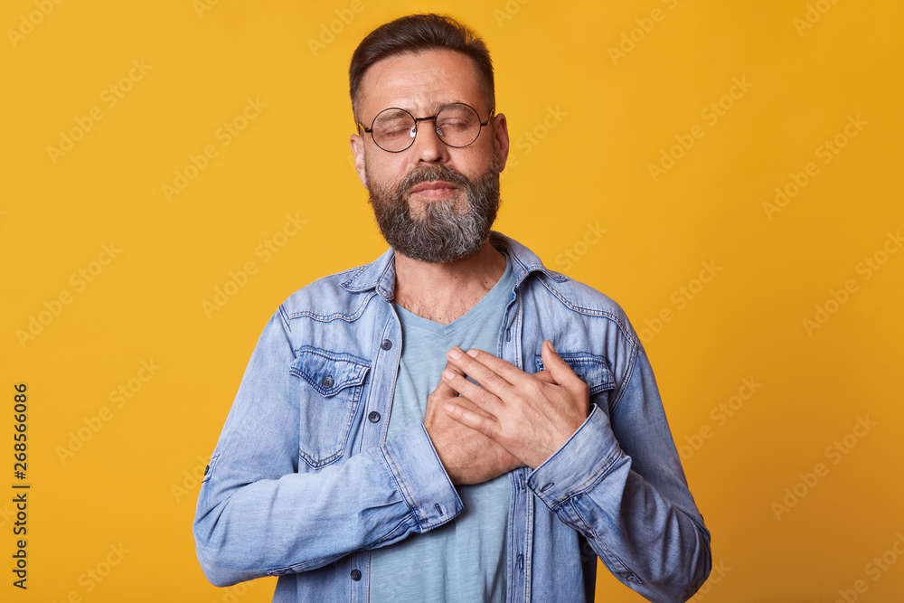Pleased bearded man with closed eyes, keeps hands on chest, dressed in fashionable denim jacket, isolated over yellow studio background, expresses his love and grateful. People emotions concept. Pleased bearded man with closed eyes, keeps hands on chest, dressed in fashionable denim jacket, isolated over yellow studio background, expresses his love and grateful. People emotions concept.