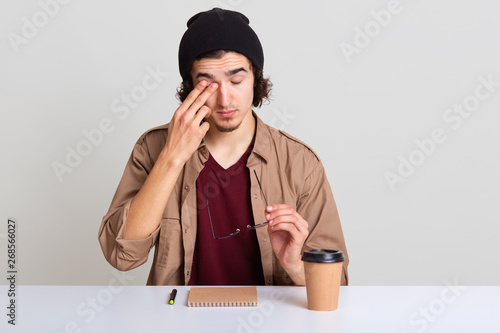Close up portrait of young unshaven male with wavy hair, sitting at desk, has pain in eyes, takes off her glasses, man wearing casually, drinks coffee and writes down something in her notebook.