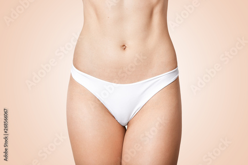 Faceless photo of woman in panties showing her perfect flat belly, lady has soft skin, wering white underwear, model posing isolated over beige background. Female health and intimate hygiene concept.