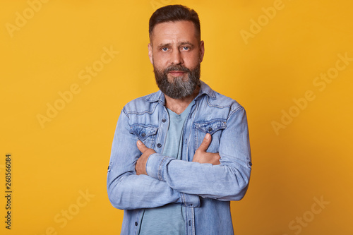 Close up portrait of handsome middle aged Caucasian man with beard, posing isolated on yellow background, wears denim jacket and gray t shirt, has serious facial expression, keeps arms crossed.