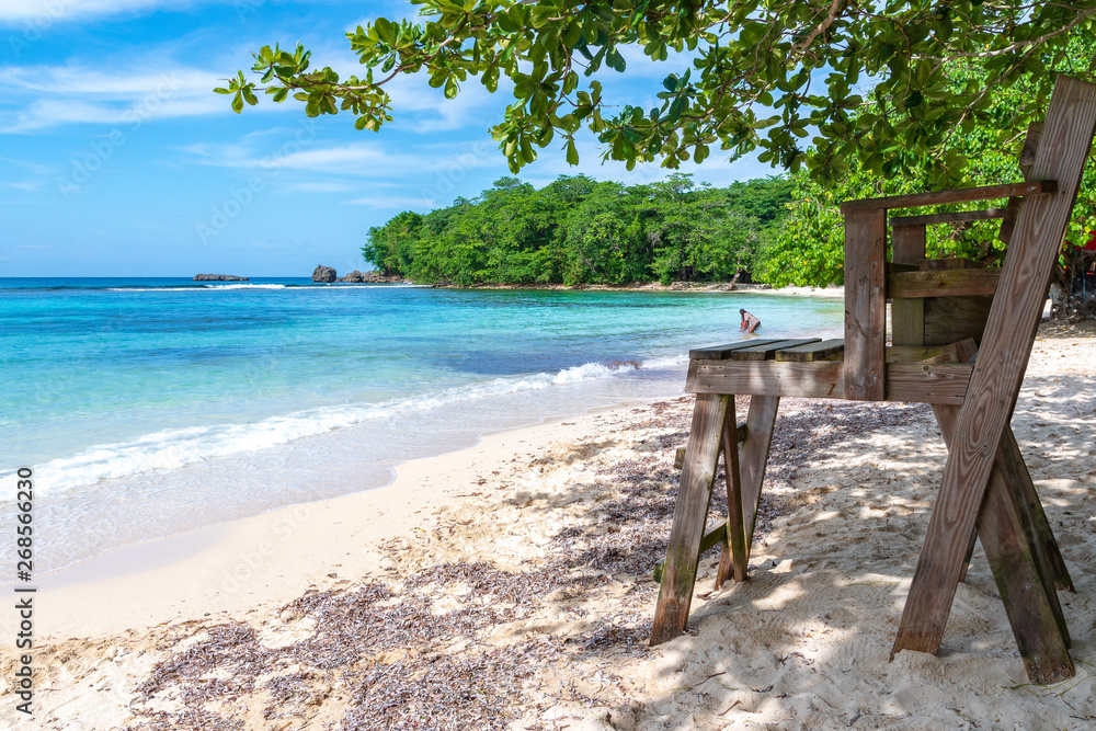Wooden lifeguard chair on beautiful white sand turquoise blue beach on tropical Caribbean island. People enjoying ocean/sea water with lush trees and large rock formations in background on summer day.