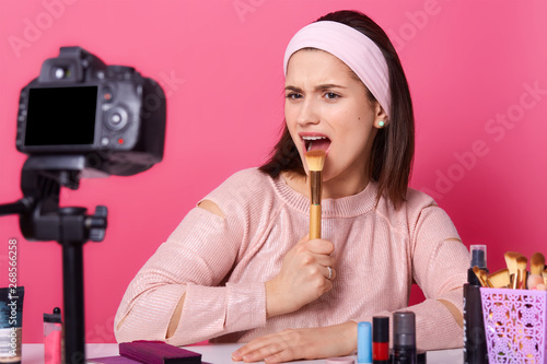 Indoor shot of charismatic artistic young blogger posing in front of camera around cosmetics, holding makeup tool in one hand, opening her mouth wide, looking directly at her camera. Shooting concept. photo