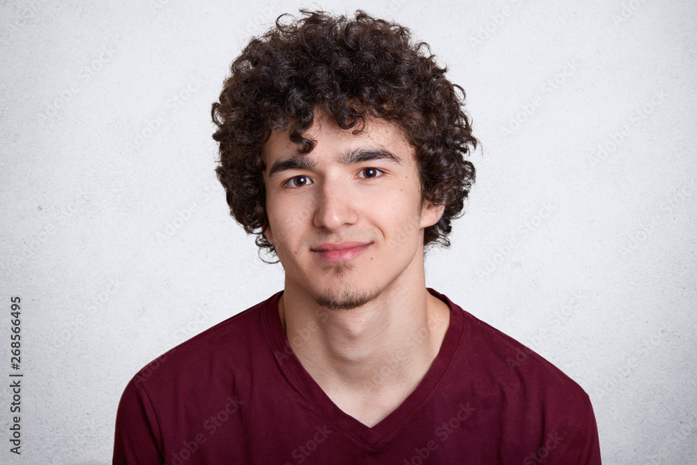 Fototapeta premium Close up portrait of sweet attractive smiling guy with curly hair and beard, having pleasant facial expression, posing isolated over white background in studio, wearing casual red sweatshirt.
