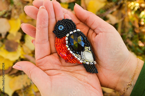 A toy bird  a handmade beaded bullfinch brooch  lying on women s palms. The background is blurred