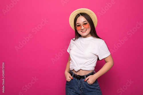 Pleased asian young woman in straw hat and sunglasses posing over pink background