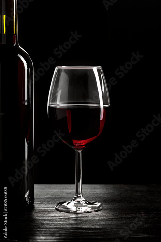 Bottle of red wine and a glass half filled with red wine, on a wooden black table, black background.