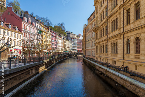Outdoor sunny view above Teplá River under shady from buildings and beautiful colourful typical European buildings with outdoor cafe along riverside in Karlovy Vary, Czech Republic.