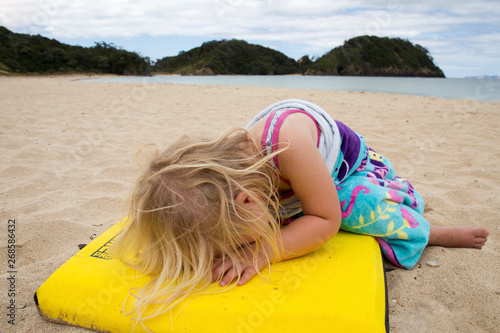 Pre school girl having a meltdown at the beach because she doesn't want to go home. Beautiful day, white sand, clear blue sea. Girl acting dramatic, tired on a yellow board with hair covering face. 