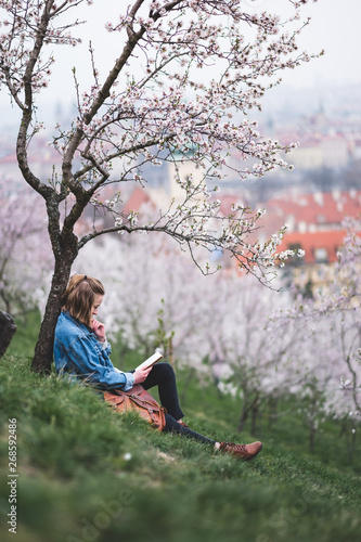 Stylish woman sitting on green grass under blossoming trees and reading a bookon hill in center of Prague, Czech Republic