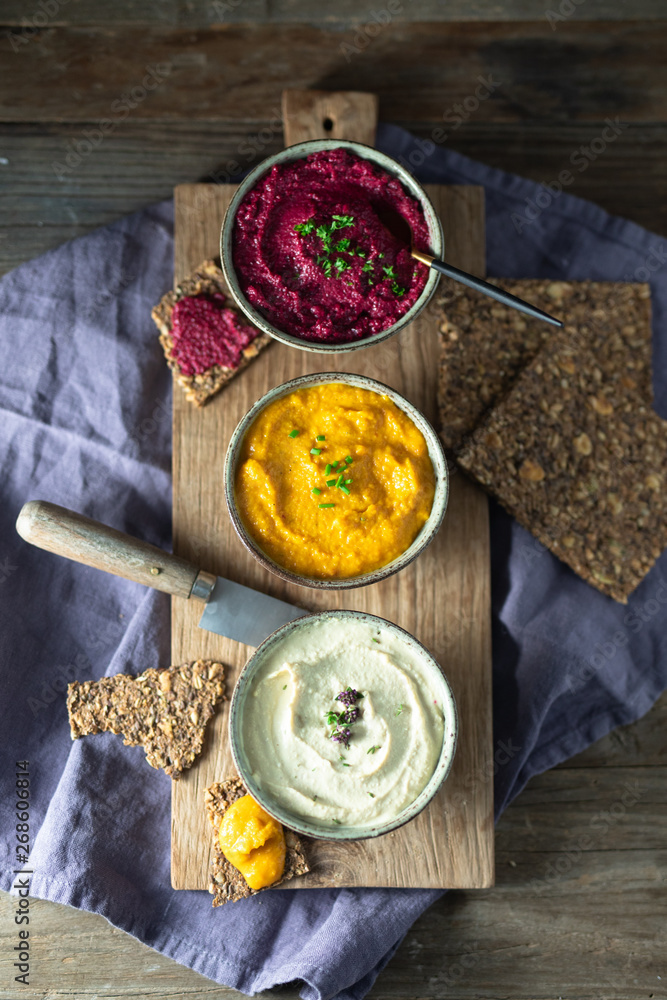 Three hummus dips, beet hummus, sweet potato or carrot or pumpkin hummus and parsnip hummus with flax seeds crackers on a rustic wooden board with a purple napkin on a dark wooden table. Top view