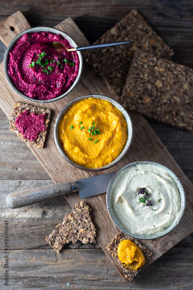 Three hummus dips, beet hummus, sweet potato or carrot or pumpkin hummus and parsnip hummus with flax seeds crackers on a rustic wooden board with a rustic knife on a dark wooden table. Top view